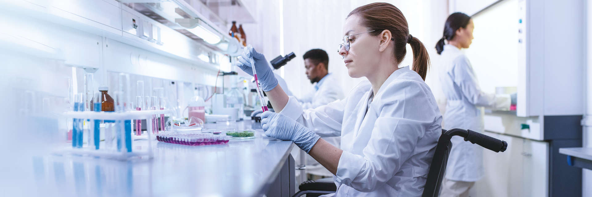 Enzymes, NGS, Caucasian female in wheelchair pipetting with a Black male and Asian female in background. Laboratory setting